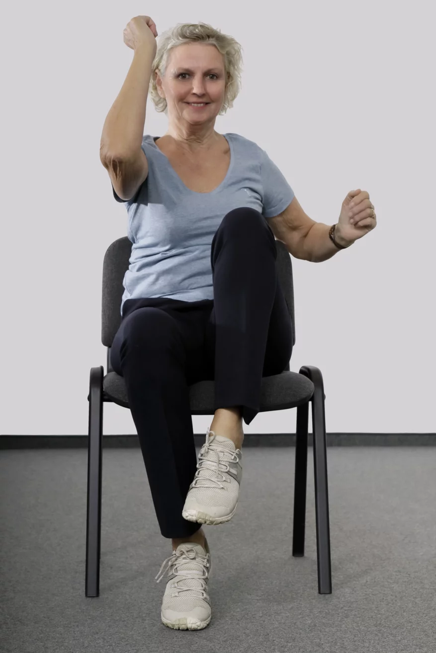 Frau sitzt auf einem Stuhl und zieht gleichzeitig das linke Knie nach oben und den angewinkelten rechten Arm