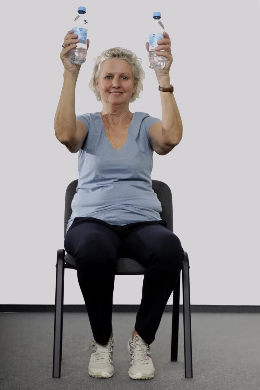Frau sitzt auf einem Stuhl, hat in jeder Hand eine kleine Wasserflasche und streckt mit angewinkelten Armen die Hände hoch