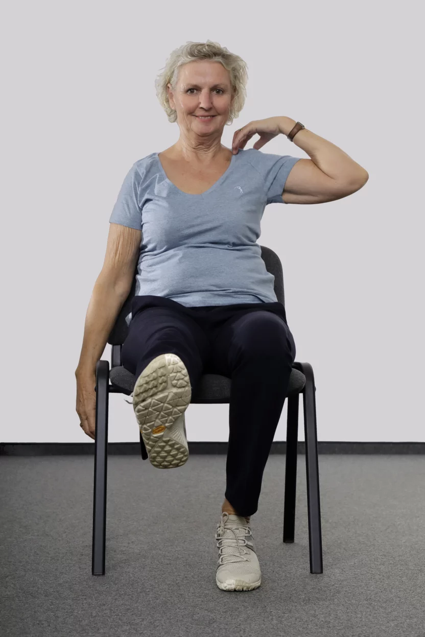 Frau sitzt auf einem Stuhl, streckt das rechte Bein aus und fasst sich mit der linken Hand an die linke Schulter